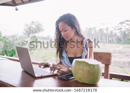 Modern Asian female blogger typing new article on laptop while sitting at table on cafe terrace and drinking coconut water 