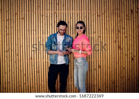 Addicted hipster influencers in streetwear clothing using modern cellphone gadgets for messaging with pda followers from social networks standing at publicity area with promotional wooden backdrop