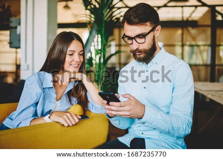 Successful female teenager looking on screen of friend's smartphone checking notification using 4G internet, hipster guy in optical spectacles sending sms message on cellular gadget via application