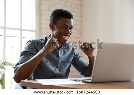 Joyful African office worker sitting at workplace desk looking at pc screen clenched fists make Yes gesture receive great offer read fantastic news, student celebrating successful exam passing concept Royalty-Free Stock Photo #1687244500