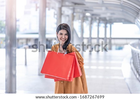beautiful asian woman holding up and showing red shopping mall bags, joyfully smiling and wearing fashionable orange long coat, within an urban district area with railing and modern platform structure