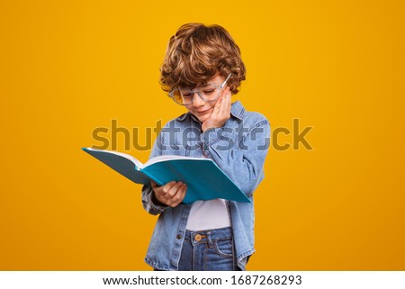 Cute kid with hand near cheek reading fascinating book during school studies against yellow background