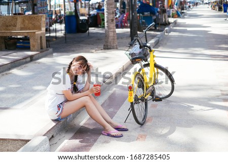 Young asian girl sitting next to her bicycle on walking shopping street close to the beach, drinking red sweet soda water from plastic cup, 