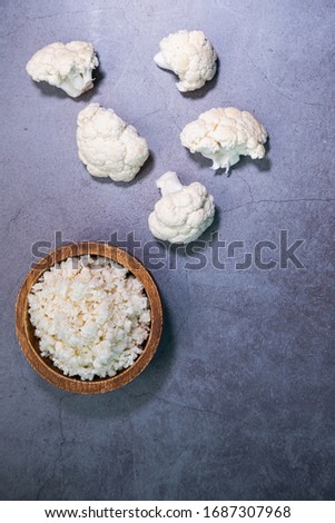 Cauliflower rice on a wooden bowl and cauliflower pieces on concrete background. Vertical picture. Top view.
