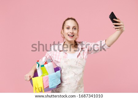Excited young woman housewife in apron hold basin with detergent bottles washing cleansers while doing housework isolated on pink background. Housekeeping concept. Doing selfie shot on mobile phone