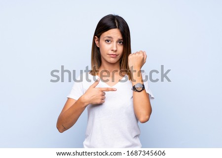 Young woman over isolated blue background showing the hand watch with serious expression