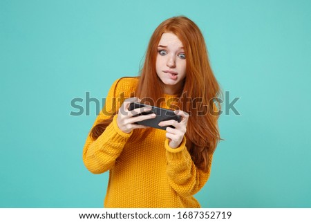 Shocked young redhead woman girl in yellow sweater posing isolated on blue turquoise background studio portrait. People lifestyle concept. Mock up copy space. Play game with mobile phone, biting lips