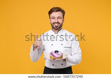 Smiling young bearded male chef cook or baker man in white uniform shirt posing isolated on yellow background in studio. Cooking food concept. Mock up copy space. Hold dessert cake showing thumb up