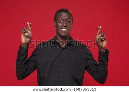 Young african american man guy in classic black shirt isolated on red background. People lifestyle concept. Mock up copy space. Wait for special moment keeping fingers crossed biting lips making wish