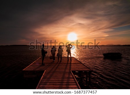 Four young guys and girls take a picture of the sunset on the water with a smart phone, from a wood pier in the coast of the Stagnone, a low sea water next to Marsala, province of Trapani, Sicily