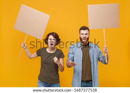 Angry protesting young two people guy girl hold in hands protest signs broadsheet blank placard on stick isolated on yellow background in studio. Protests strikes pickets concept. Youth against city