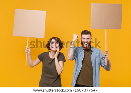 Joyful protesting two people guy girl hold protest signs broadsheet blank placard on stick doing winner gesture isolated on yellow background. Protests strikes pickets concept. Youth against city