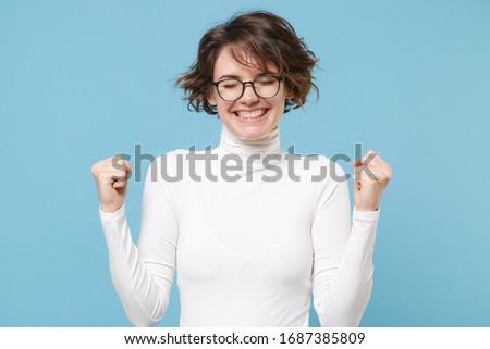 Smiling young brunette woman girl in casual white clothes, eyeglasses posing isolated on pastel blue background. People lifestyle concept. Mock up copy space. Doing winner gesture keeping eyes closed