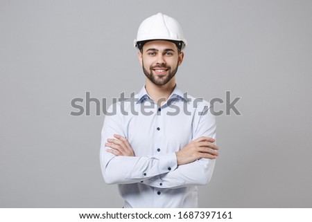 Smiling young unshaven business man in light shirt protective construction helmet isolated on grey background. Achievement career wealth business concept. Mock up copy space. Holding hands crossed Royalty-Free Stock Photo #1687397161