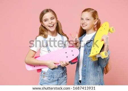 Funny little kids girls 12-13 years old in white t-shirt, denim clothes isolated on pastel pink wall background. Childhood lifestyle concept. Mock up copy space. Hold skateboards showing victory sign