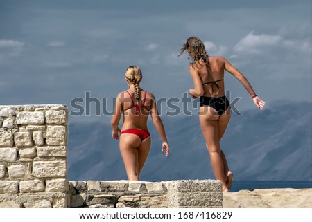 Two beautiful girls in bikini on sea beach with mountains and blue sky on blurred background and stone ancient wall on foreground on Corfu island in Greece