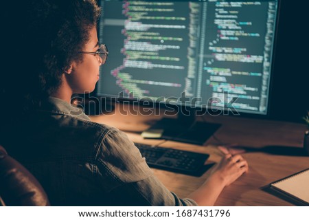 Back behind view photo of dark skin programmer lady look big monitor check id-address work overtime check debugging system wear specs casual shirt sit table late night office indoors