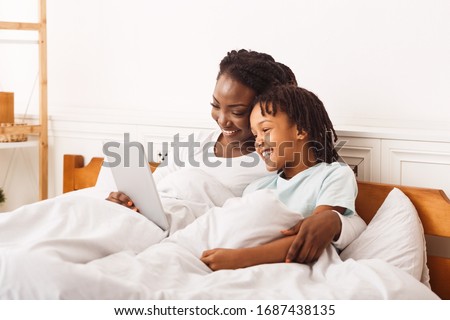 Happy Loving Family Concept. African mother and her daughter lying in bed, having fun with digital tablet, copyspace