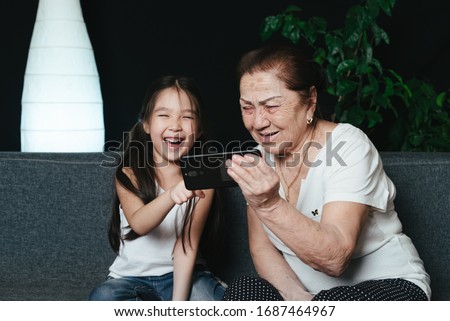 Asian gandmother with granddaughter sitting on a sofa watching funny video from smartphone