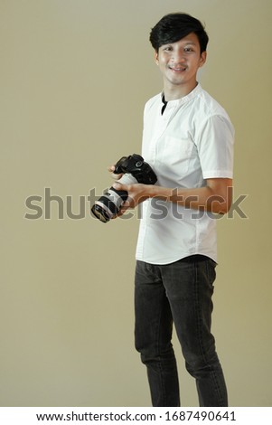 Portrait of asian man in white shirt holding digital camera with smile. Handsome male photographer standing isolated on plain background copy space. Photography business concept.
