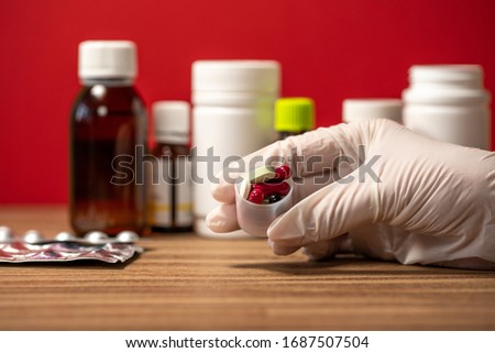 Closeup view photography of gloved hand of white woman holding small plastic container full of different tablets, drugs, vitamins isolated at blurry defocused medical jars and bottles background.