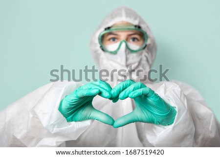 Medical worker in safety glasses, mask and suit makes a heart sign. Green homogeneous background. Copy space. Confronting coronavirus.