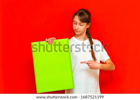 Surprised caucasian young long haired girl holding and poke at bright green folder with a banner advertisement on a red studio background. copy space banner
