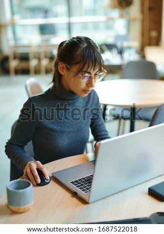 Young woman in eyeglasses concentrating on screen and typing on laptop while sitting at desk at workplace or cafe. Concept remote work, freelance, using laptop computer.