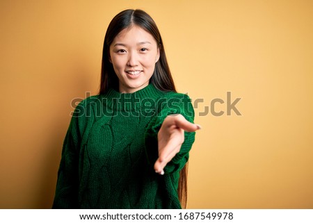 Young beautiful asian woman wearing green winter sweater over yellow isolated background smiling cheerful offering palm hand giving assistance and acceptance.