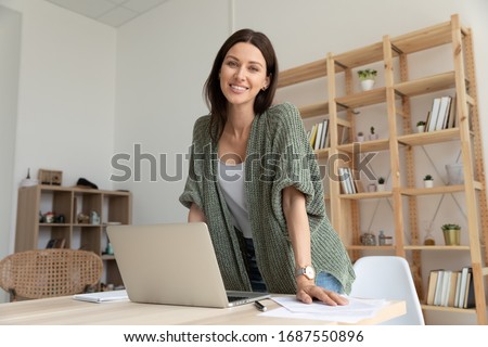 Portrait of smiling young businesswoman look at camera posing at workplace in modern office, happy Caucasian female employee working with documents using modern laptop gadget, career success concept