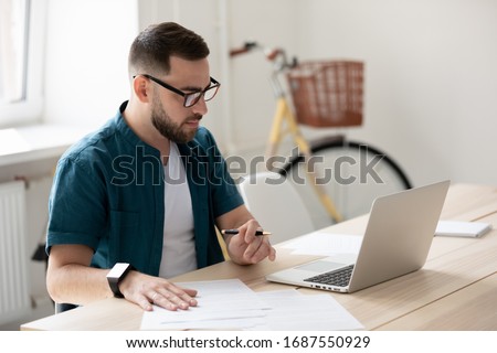 Focused young male employee in glasses watch webinar on laptop make notes, concentrated millennial man worker sit at desk in office study or work on computer, handwrite on paper summarizing Royalty-Free Stock Photo #1687550929