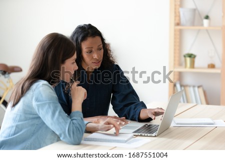 Focused diverse female colleagues sit at desk in office brainstorm discuss business idea use laptop, concentrated woman coworkers look at computer screen work together at meeting, cooperation concept Royalty-Free Stock Photo #1687551004