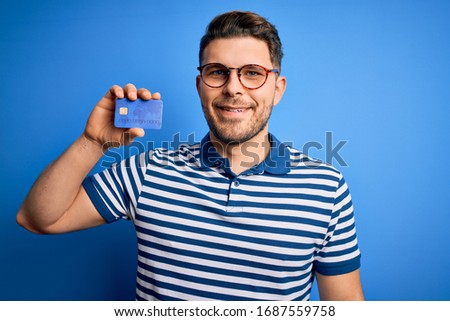 Young man with blue eyes wearing glasses and holding credit card over blue background with a happy face standing and smiling with a confident smile showing teeth