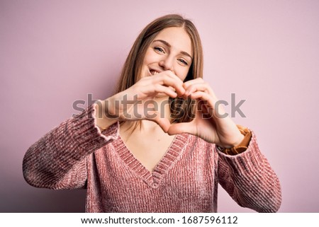 Young beautiful redhead woman wearing casual sweater over isolated pink background smiling in love doing heart symbol shape with hands. Romantic concept.