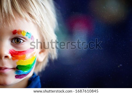 Beautiful blond toddler boy with rainbow painted on his face and messy hands, smiling happily