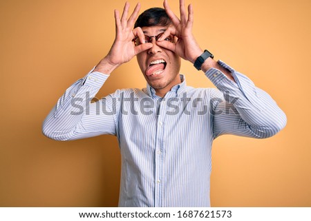 Young handsome hispanic business man wearing nerd glasses over yellow background doing ok gesture like binoculars sticking tongue out, eyes looking through fingers. Crazy expression.
