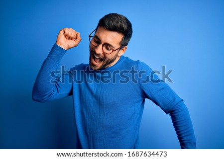 Young handsome man with beard wearing casual sweater and glasses over blue background Dancing happy and cheerful, smiling moving casual and confident listening to music