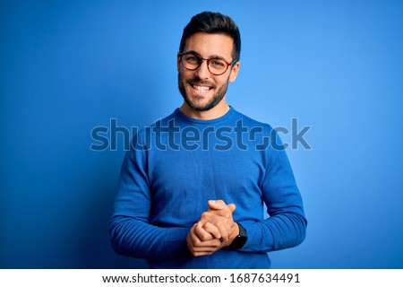 Young handsome man with beard wearing casual sweater and glasses over blue background with hands together and crossed fingers smiling relaxed and cheerful. Success and optimistic Royalty-Free Stock Photo #1687634491