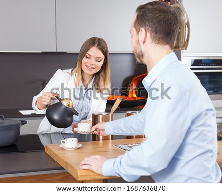 Smiling attractive girl pouring tea into cup to boyfriend in stylish home kitchen
