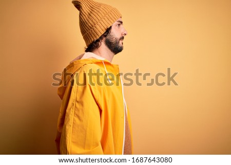 Young handsome man with beard wearing raincoat for rainy day over yellow background looking to side, relax profile pose with natural face with confident smile.
