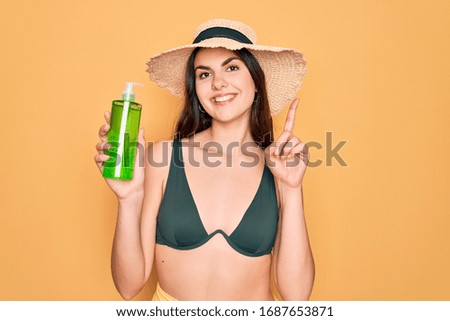 Young beautiful girl wearing swimwear bikini and summer hat holding alove vera sun protection surprised with an idea or question pointing finger with happy face, number one