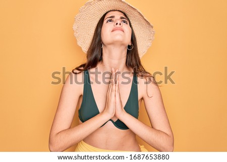 Young beautiful girl wearing swimwear bikini and summer sun hat over yellow background begging and praying with hands together with hope expression on face very emotional and worried. Begging.