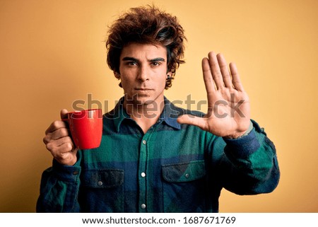 Young handsome man drinking cup of coffee standing over isolated yellow background with open hand doing stop sign with serious and confident expression, defense gesture