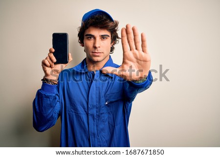 Young mechanic man wearing uniform holding smartphone over isolated white background with open hand doing stop sign with serious and confident expression, defense gesture