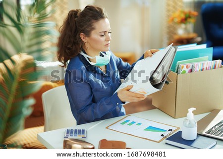 trendy female in blue blouse with medical mask organising a temporary home office during the coronavirus epidemic in the modern house in sunny day. Royalty-Free Stock Photo #1687698481