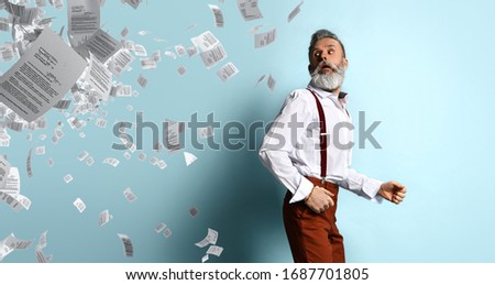 Gray-haired, aged man in white shirt, brown pants and suspenders, bracelet. He running from sheets of paper or documents randomly located on left side of photo, blue background. Close up, copy space