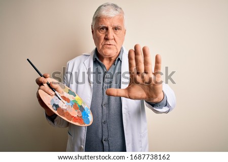 Senior handsome hoary painter man painting using brush and palette with colors with open hand doing stop sign with serious and confident expression, defense gesture
