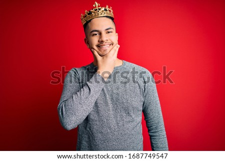 Young handsome man wearing golden crown of prince over isolated red background looking confident at the camera smiling with crossed arms and hand raised on chin. Thinking positive.