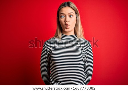 Young beautiful blonde woman wearing casual striped sweater over red isolated background making fish face with lips, crazy and comical gesture. Funny expression.