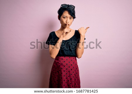 Young beautiful asian girl wearing casual dress standing over isolated pink background asking to be quiet with finger on lips pointing with hand to the side. Silence and secret concept.
